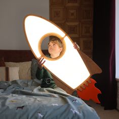 a young boy sitting on top of a bed holding a large light up rocket ship