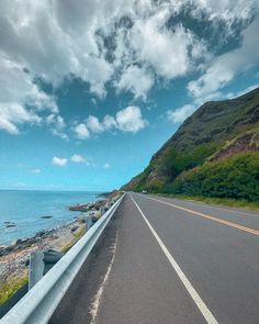 an empty highway with the ocean and mountains in the background, under a cloudy blue sky