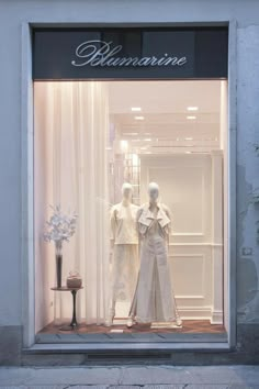 two mannequins in front of a store window with white curtains and flowers