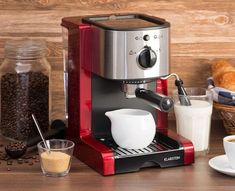 a red espresso machine sitting on top of a wooden table next to a basket of croissants