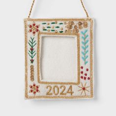 a white frame hanging from a rope with embroidered flowers and leaves on the front, decorated in gold thread