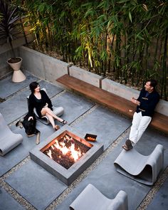 two people sitting around a fire pit in the middle of an outdoor area with chairs and tables