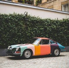 a multicolored porsche sits parked in front of a wall with green bushes behind it