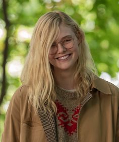 a woman with long blonde hair and glasses smiles at the camera while standing in front of trees