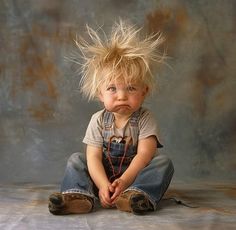 a young child with blonde hair sitting on the ground wearing overalls and looking at the camera