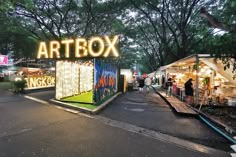 an artbox is lit up at night in the middle of a park with people walking around