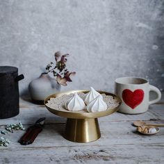 two small white desserts on a gold plate next to coffee cups and spoons