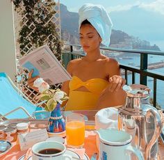 a woman sitting at a table reading a newspaper and drinking orange juice in front of the ocean