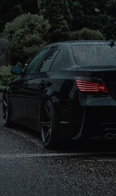 the back end of a black car parked in a parking lot with trees behind it