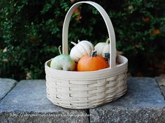 a white basket filled with pumpkins sitting on top of a stone wall next to a bush