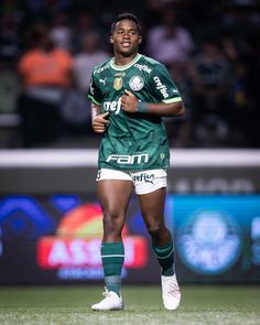 a soccer player in green and white uniform running on the field with his hands behind his back