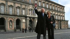 a man and woman standing in front of an old building waving at the camera with their hands up