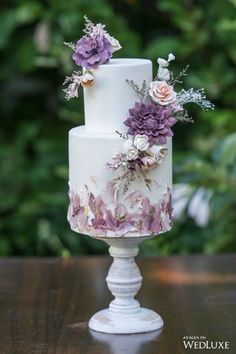 a white and purple wedding cake with flowers on the top is sitting on a table