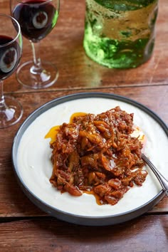 a white plate topped with meat and sauce next to two glasses of wine on a wooden table