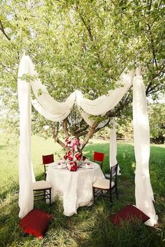 a table set up for a wedding under a tree with white draping and red chairs