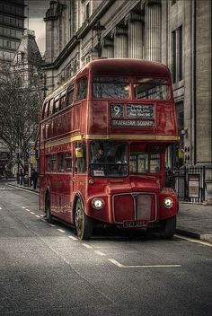 a red double decker bus driving down a street next to tall buildings on either side