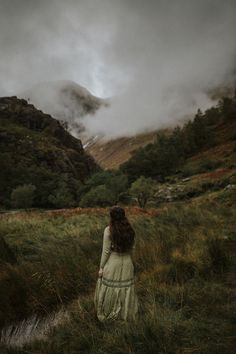 a woman standing in the middle of a lush green field with mountains in the background