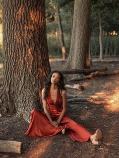 a woman in a red dress sitting on the ground next to a tree and looking up