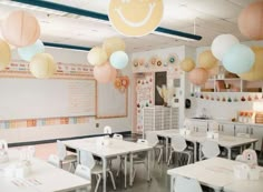 a classroom filled with lots of white tables and chairs covered in paper lanterns hanging from the ceiling