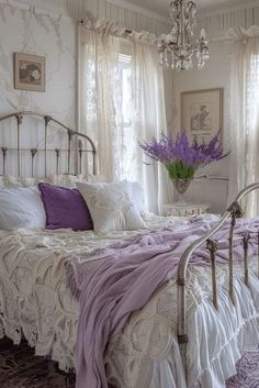 a white bed topped with purple pillows next to a chandelier and window covered in curtains