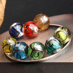 seven colorful glass beads sitting on top of a metal tray next to an orange leaf