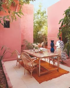 an outdoor dining area with pink walls and wooden table surrounded by wicker chairs, potted plants, and rug