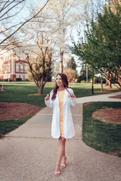 a woman standing on a sidewalk wearing a white coat and yellow dress with her hands in her pockets