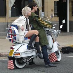 a man and woman riding on the back of a white scooter down a street