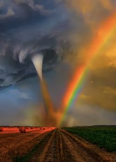 a rainbow appears in the sky over a dirt road and farm field with silos