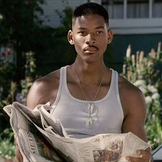 a man in tank top holding a newspaper and looking at the camera while standing next to a car