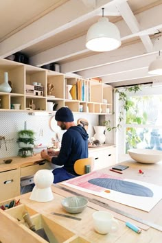 a man sitting at a table working on some art work in a room with lots of shelves