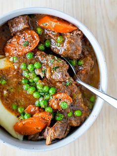 a bowl filled with meat, peas and potatoes on top of a wooden table next to a fork