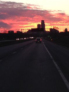 a car driving down the road in front of an orange and pink sky at sunset