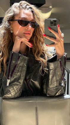 a woman in black jacket and sunglasses taking a selfie with her cell phone while sitting on a chair