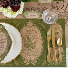 a place setting on a table with goldware and green floral napkins in the center