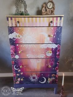 a painted chest of drawers with an image of the sun and moon on it, next to a candle