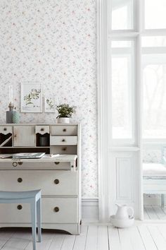 a white desk sitting in front of a window next to a chair and vase on top of a table