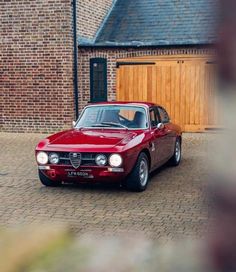 a red alfa coupe parked in front of a brick building with two garage doors on the side