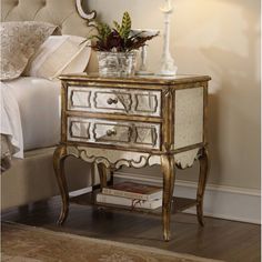 a white and gold mirrored nightstand with flowers on top, next to a bed in a bedroom