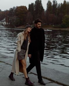 a man and woman walking next to each other on the sidewalk near water with trees in the background