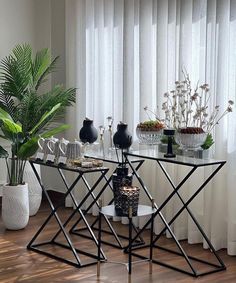 two tables with vases and plants on them in front of a window covered by white drapes