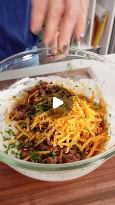 a person mixing ingredients in a glass bowl on a wooden table with cheese and other items