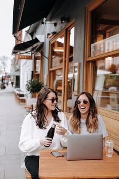 two women sitting at a table with a laptop and drink in front of their faces