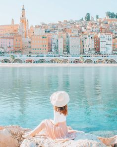 a woman sitting on top of a rock next to the ocean with buildings in the background