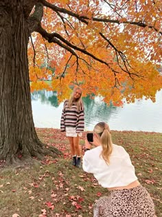 two women sitting under a tree in front of a lake with leaves on the ground