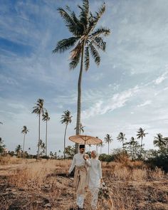 two people standing under an umbrella in the middle of a field with palm trees behind them