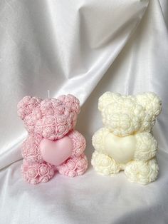 two small teddy bears sitting next to each other on a white cloth covered surface with a heart shaped candle in the middle