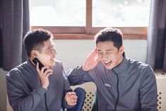 two young men sitting at a table talking on cell phones while one holds his hand up to his ear