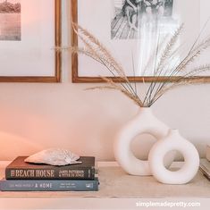 two framed pictures hang on the wall above a white vase with dried grass in it