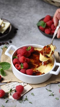 a person holding a spoon full of food with raspberries on the table next to it